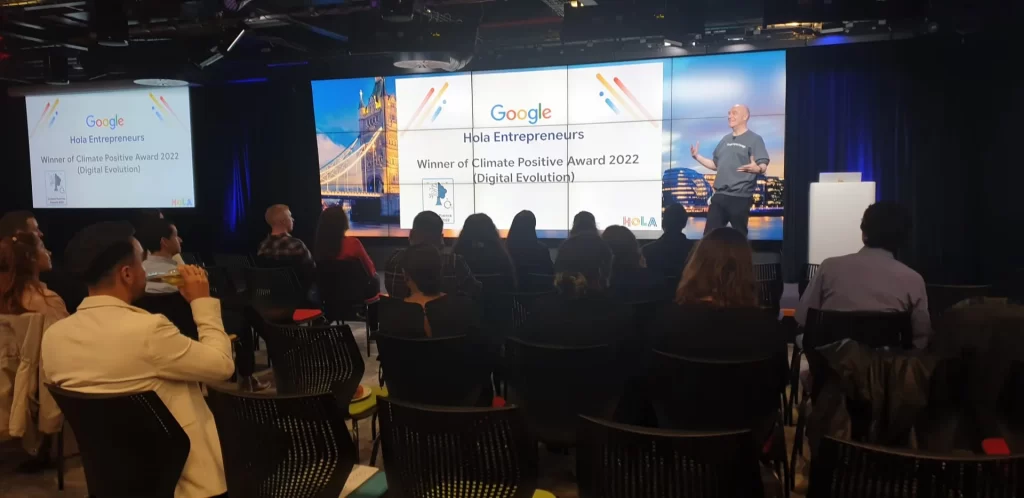 Glenn White speaking on stage at the Google Hola Entrepreneurs event in London, with an audience watching and a screen showing the Climate Positive Award for Digital Evolution 2022.