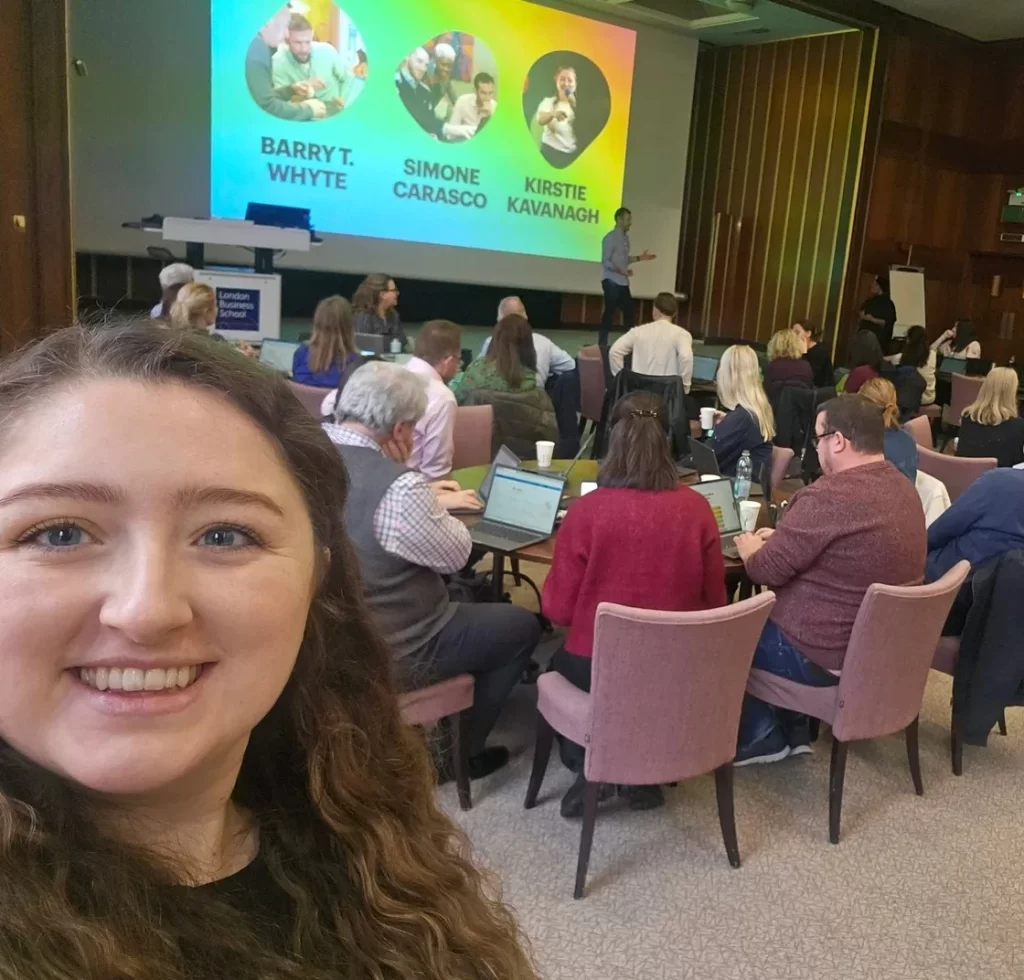 Kirstie Kavanagh from City Folk Digital smiling in the foreground of an AI training workshop at London Business School, with attendees working on laptops and a presentation screen showing speaker names.
